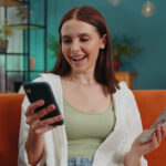 Smiling happy young woman at home counting money cash use smartphone, income, saves, lottery win