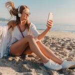 young attractive blond smiling woman taking selfie photo on phone on vacation sitting on beach in headphones listening to music relaxed