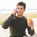 Young caucasian man at outdoors using mobile phone and holding a coffee